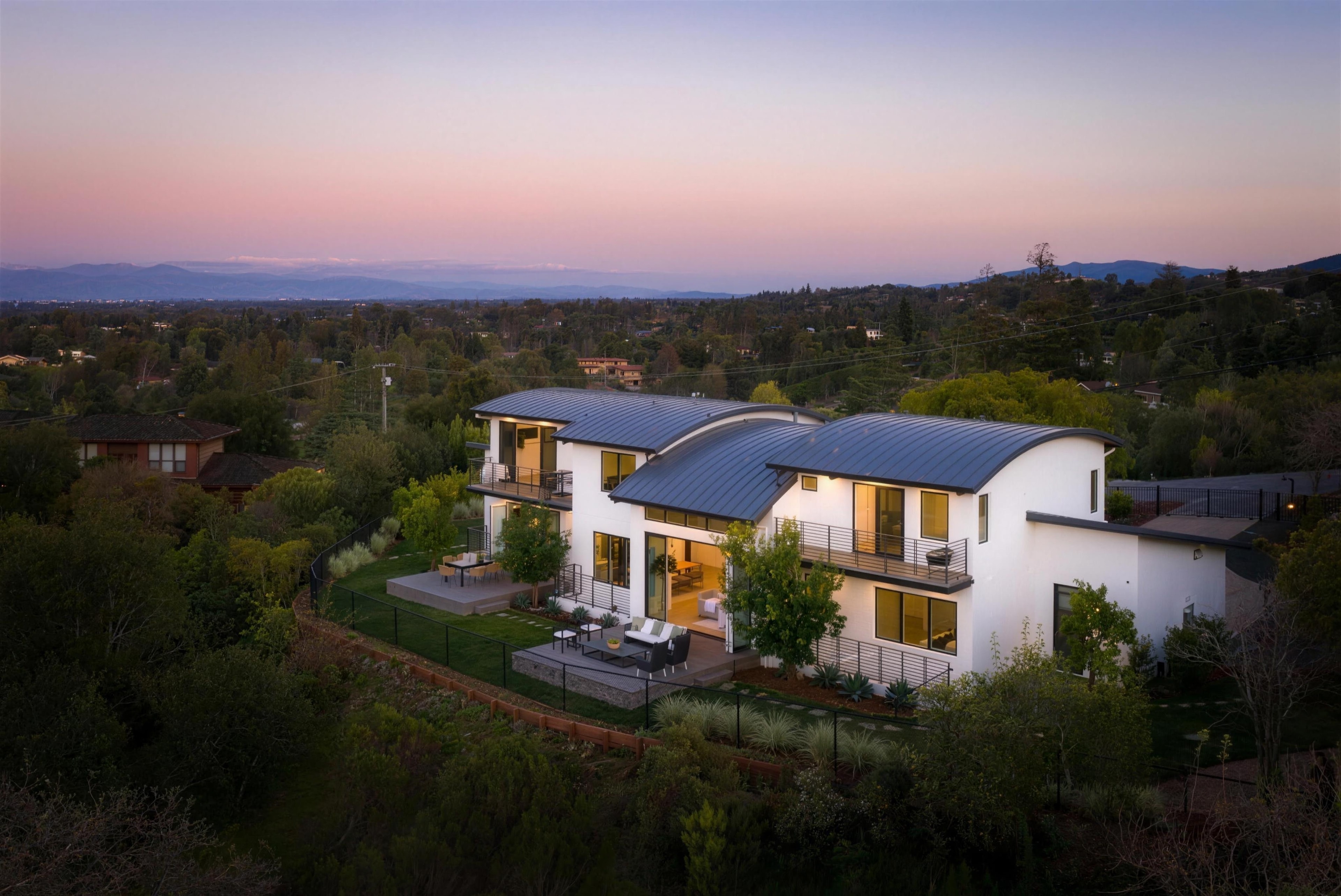 An aerial view at dusk of a white two-storey house with curved metal roofs, lit from within, on a wooded hillside above a valley under a pink sky
